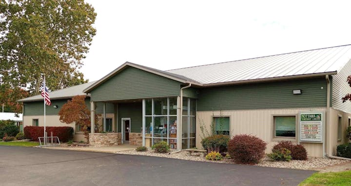 A library building with stone and green siding, flagpole, plants, and a sign reading "Nottawa Township Library" with hours listed.
