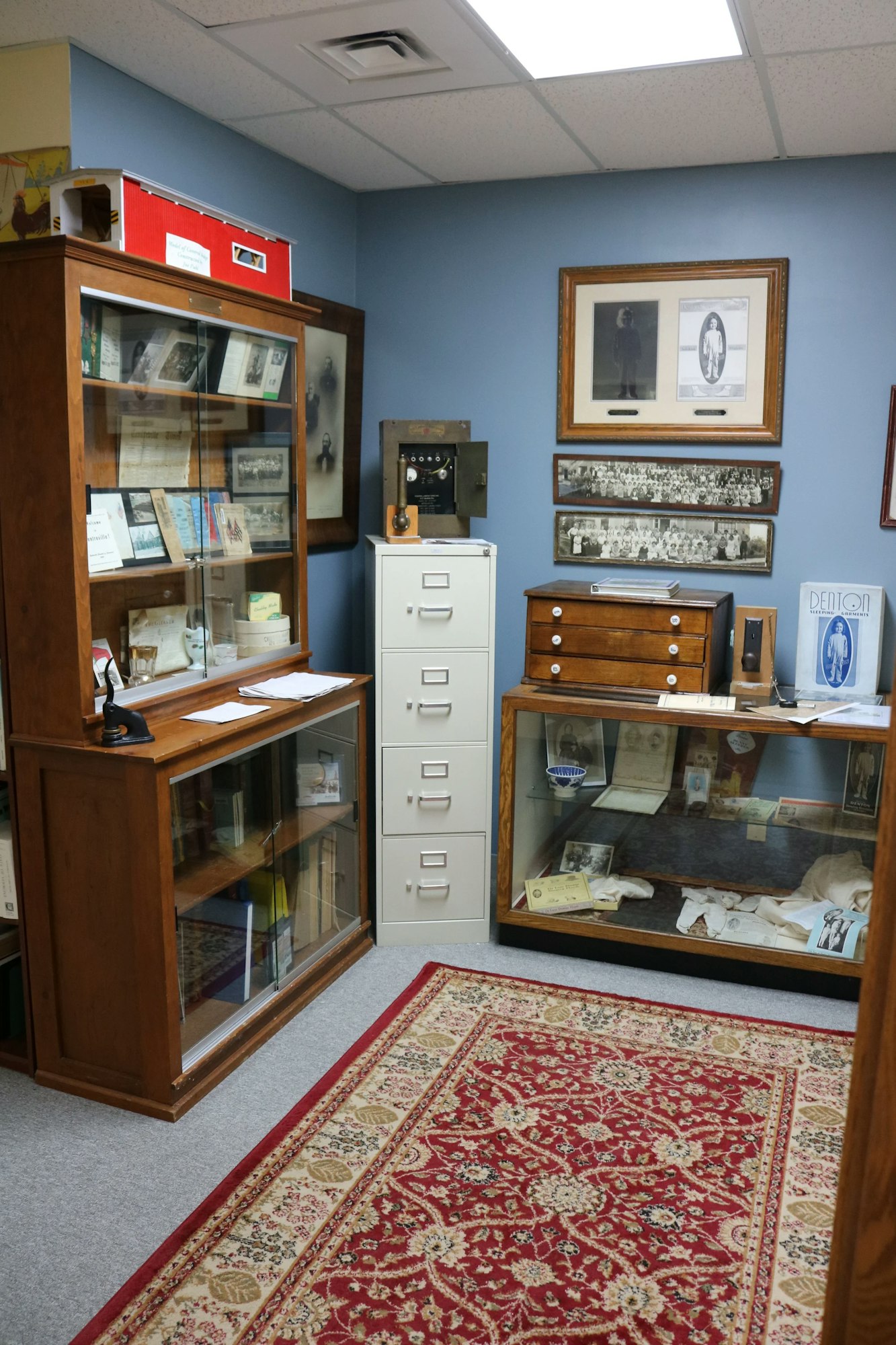 A room with glass cabinets displaying old photos and documents, a filing cabinet, vintage artifacts, and a patterned red rug.