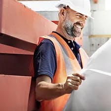 A smiling worker with a hard hat and reflective vest, reviewing a large paper plan.