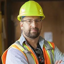 A man with a beard wearing a yellow hard hat, safety vest, and glasses.
