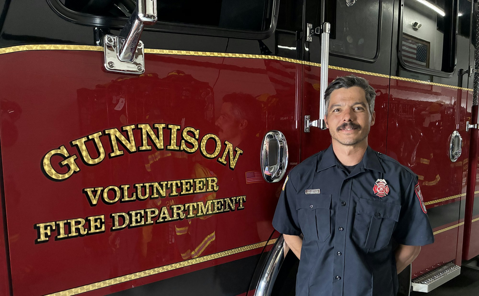 A firefighter stands beside a fire truck labeled "Gunnison Volunteer Fire Department."