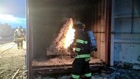 A firefighter is near a burning pile inside a training structure, with another firefighter in the background.