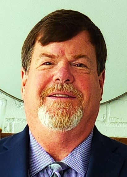 A person with a beard, wearing a suit and tie, against a light background.