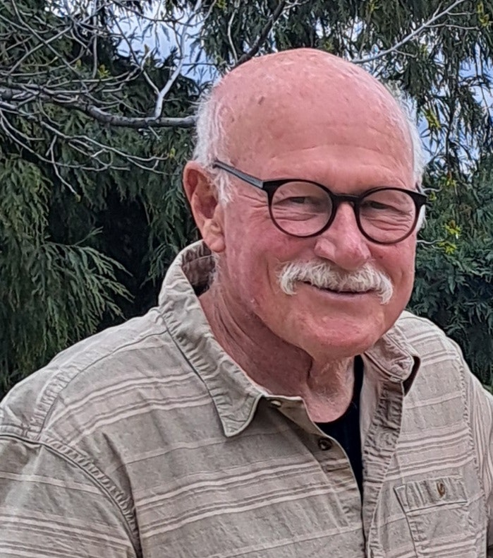 A smiling older man with glasses and a mustache stands outdoors, surrounded by greenery.