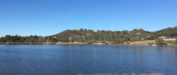 A serene lake view surrounded by hills and trees under a clear blue sky, featuring houses scattered along the shore.