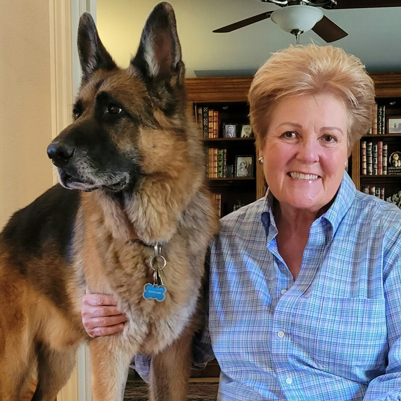 A person smiling next to a German Shepherd in a room with bookshelves and a ceiling fan.