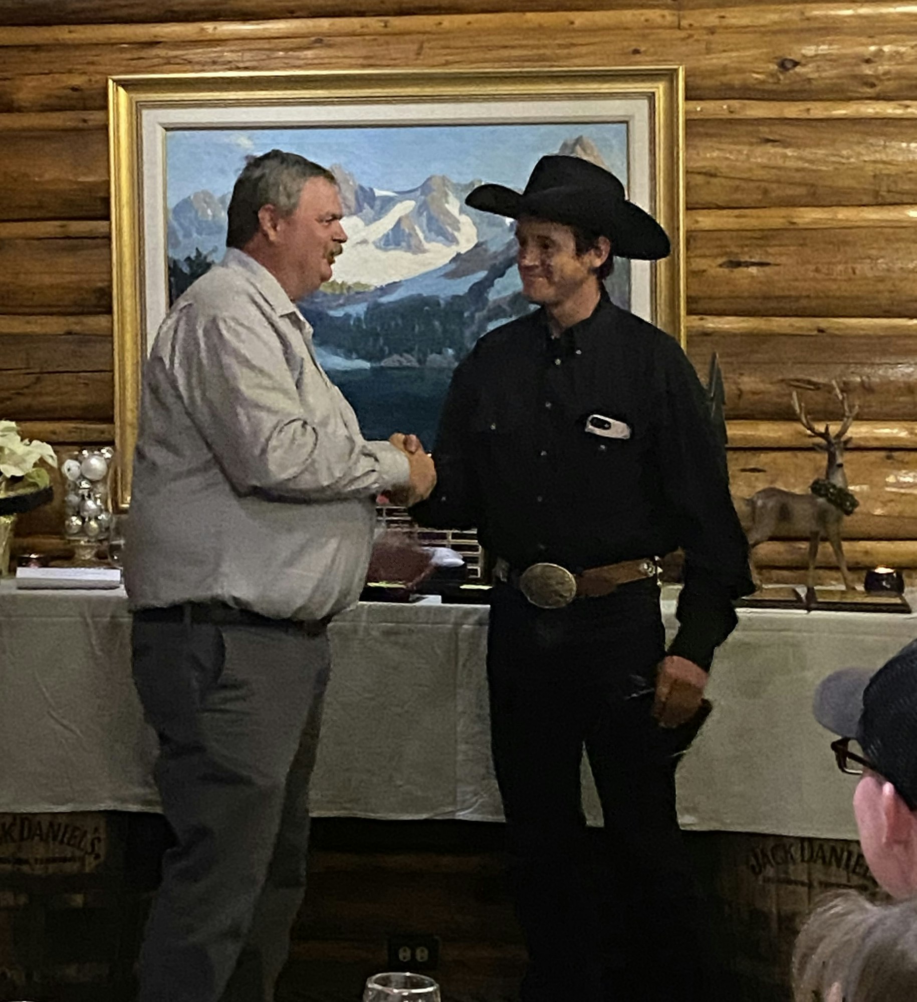 Two men shaking hands, one in cowboy attire, with a landscape painting and trophy buckles in the background.