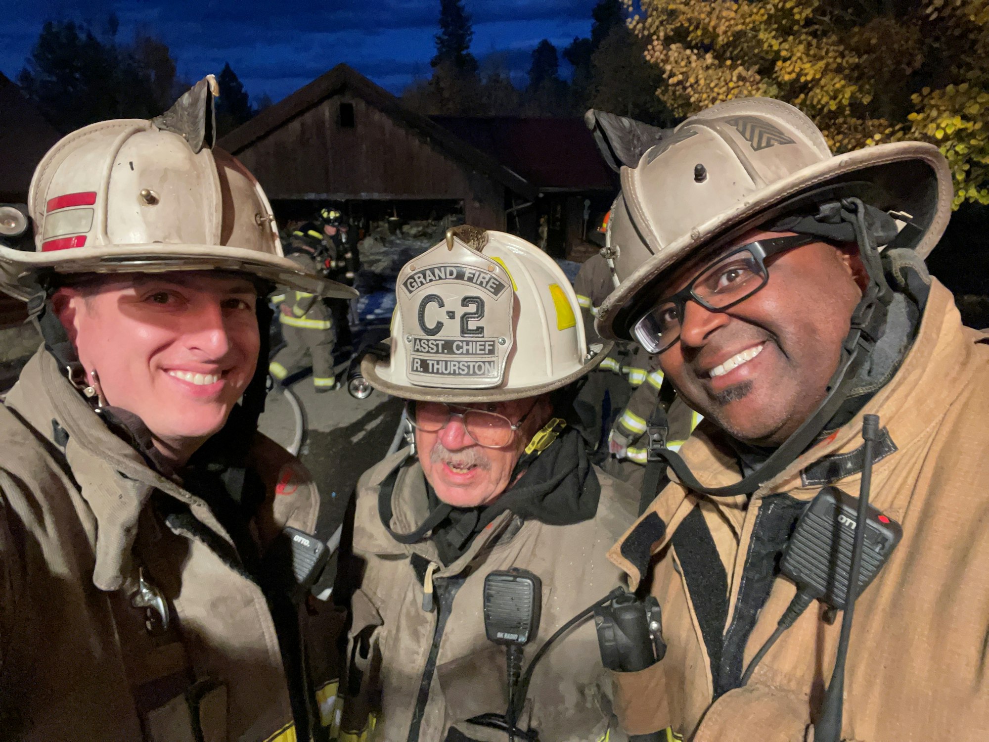 Three firefighters posing for a selfie, smiling, with gear on, and a hazy outdoor background.