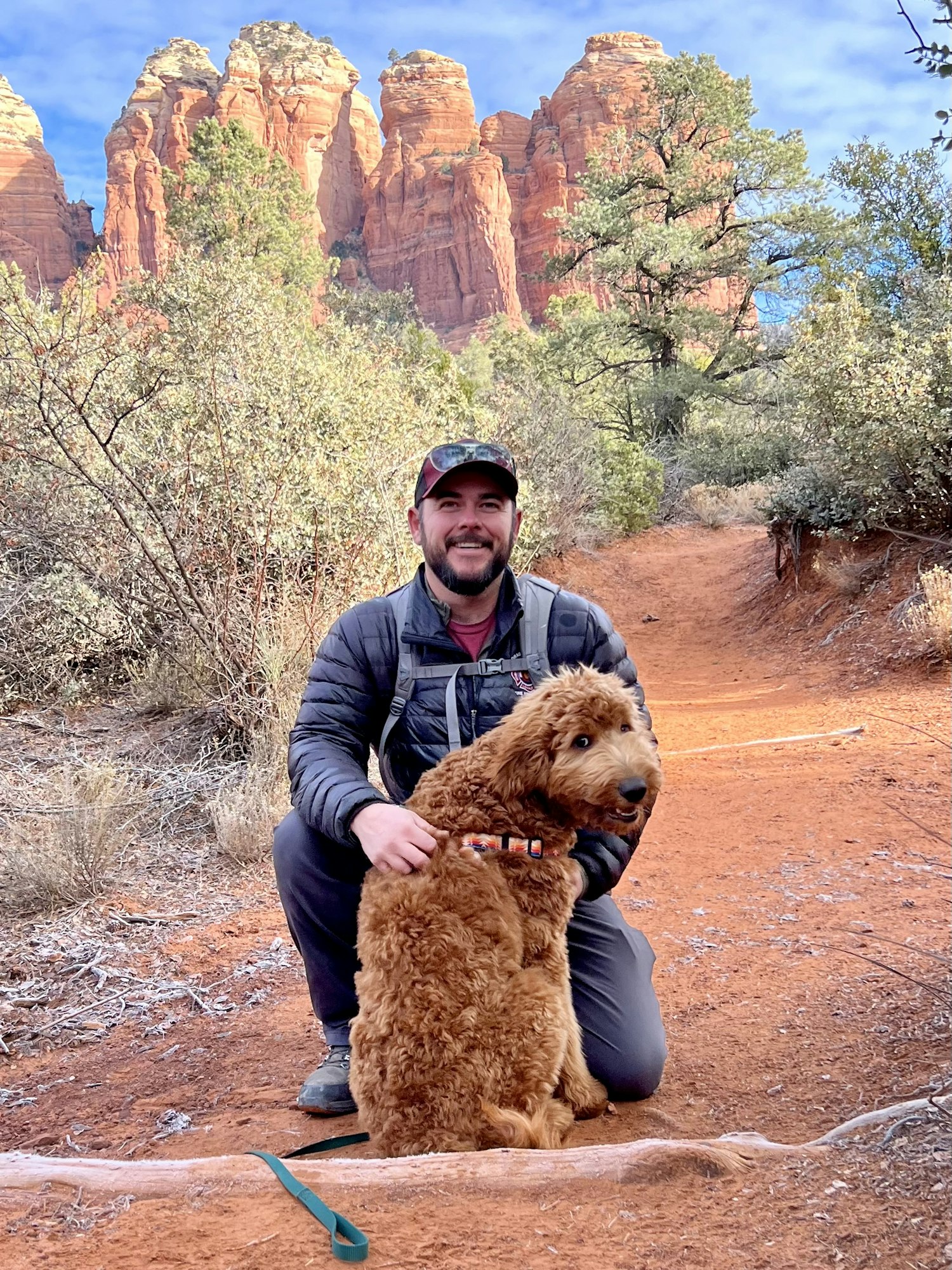 A smiling person and a fluffy dog outdoors with red rock formations in the background.