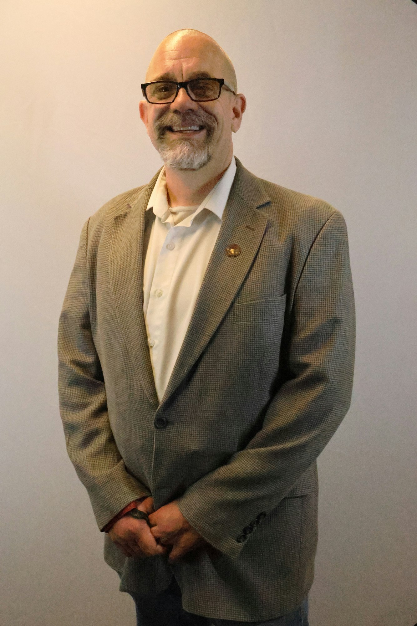 A person wearing glasses and a suit jacket, smiling, standing against a plain background.