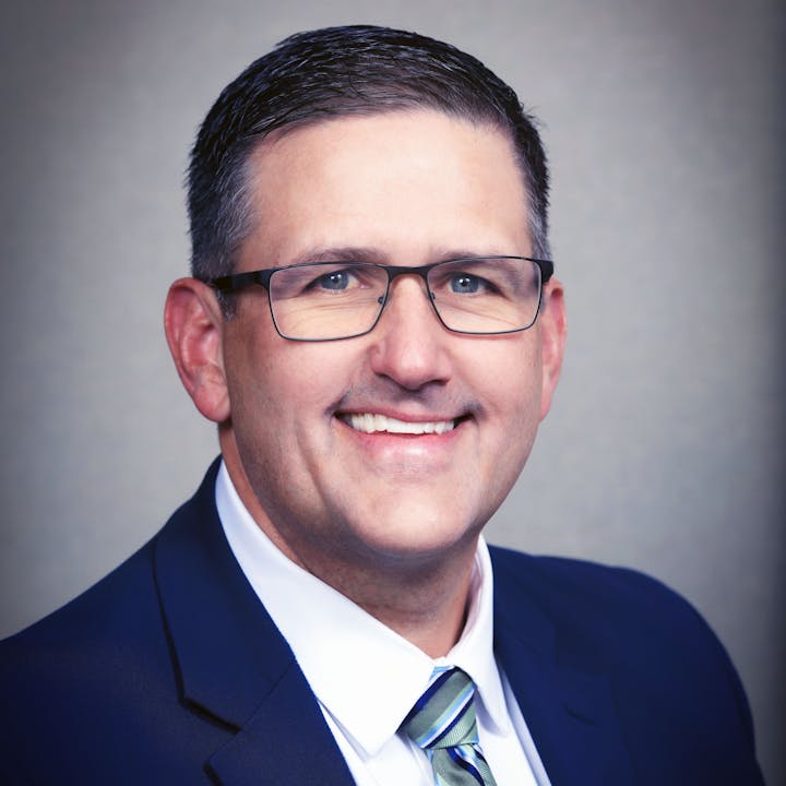 A smiling person wearing glasses, a suit, and a tie against a neutral background.