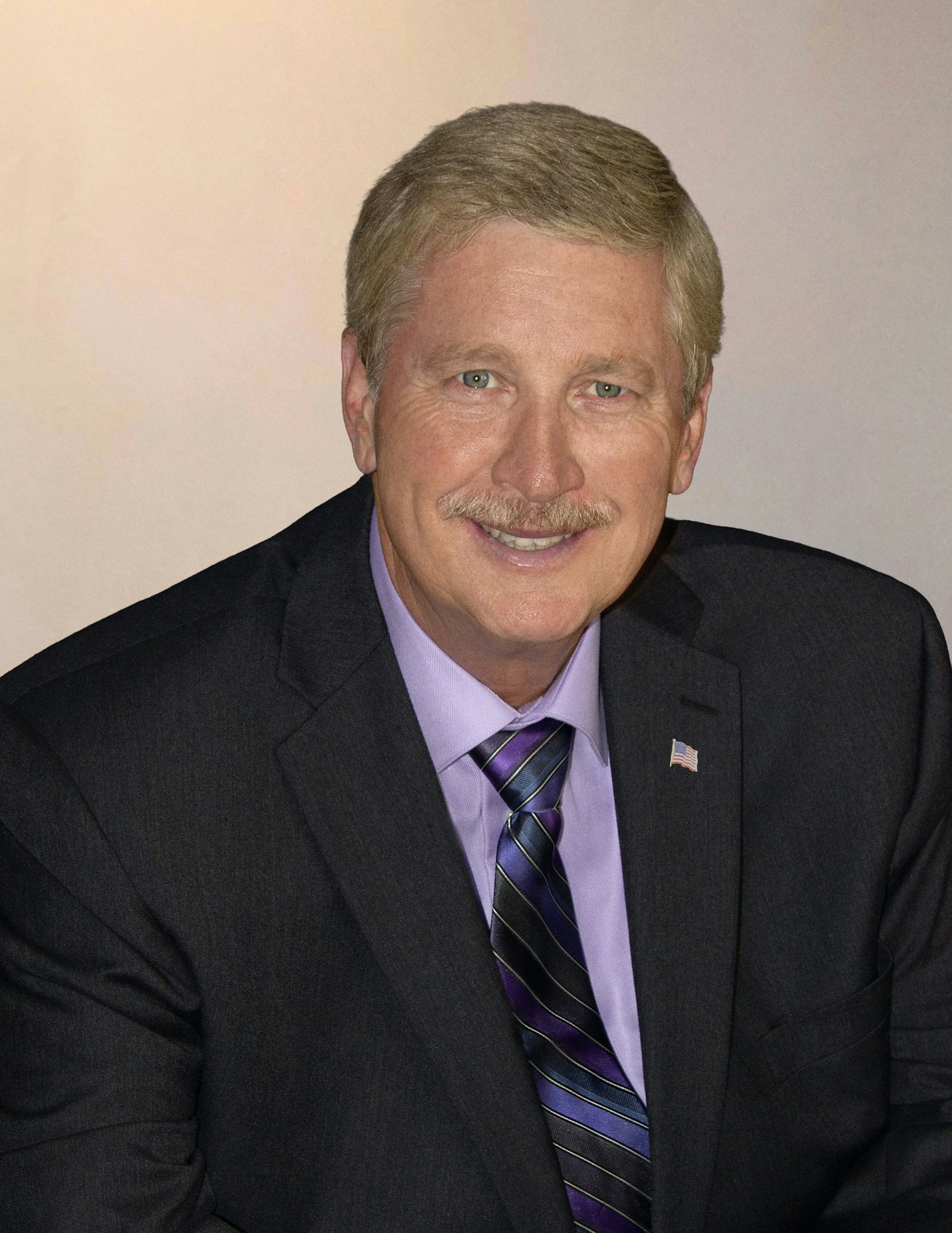 A man with a mustache in a suit and tie, smiling at the camera.