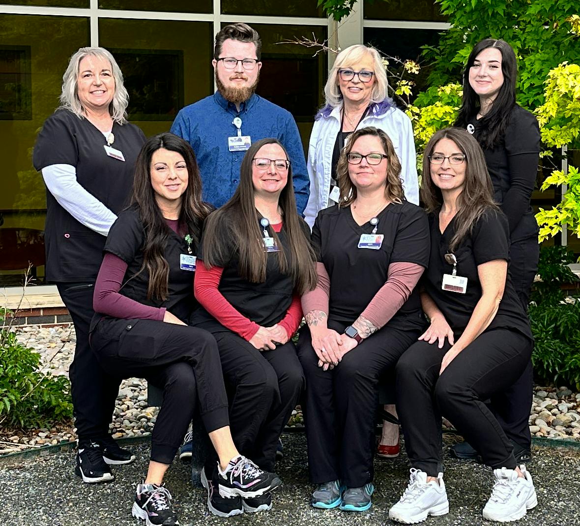 A group of nine people posing outside, some in uniforms with name tags, standing and kneeling in front of a building with greenery.