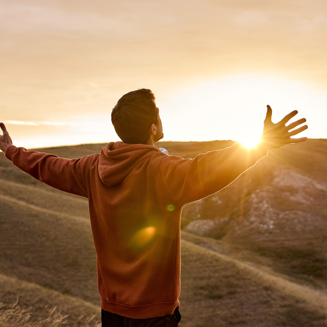 A person stands with arms outstretched, embracing a sunrise over rolling hills, evoking feelings of freedom and connection to nature.