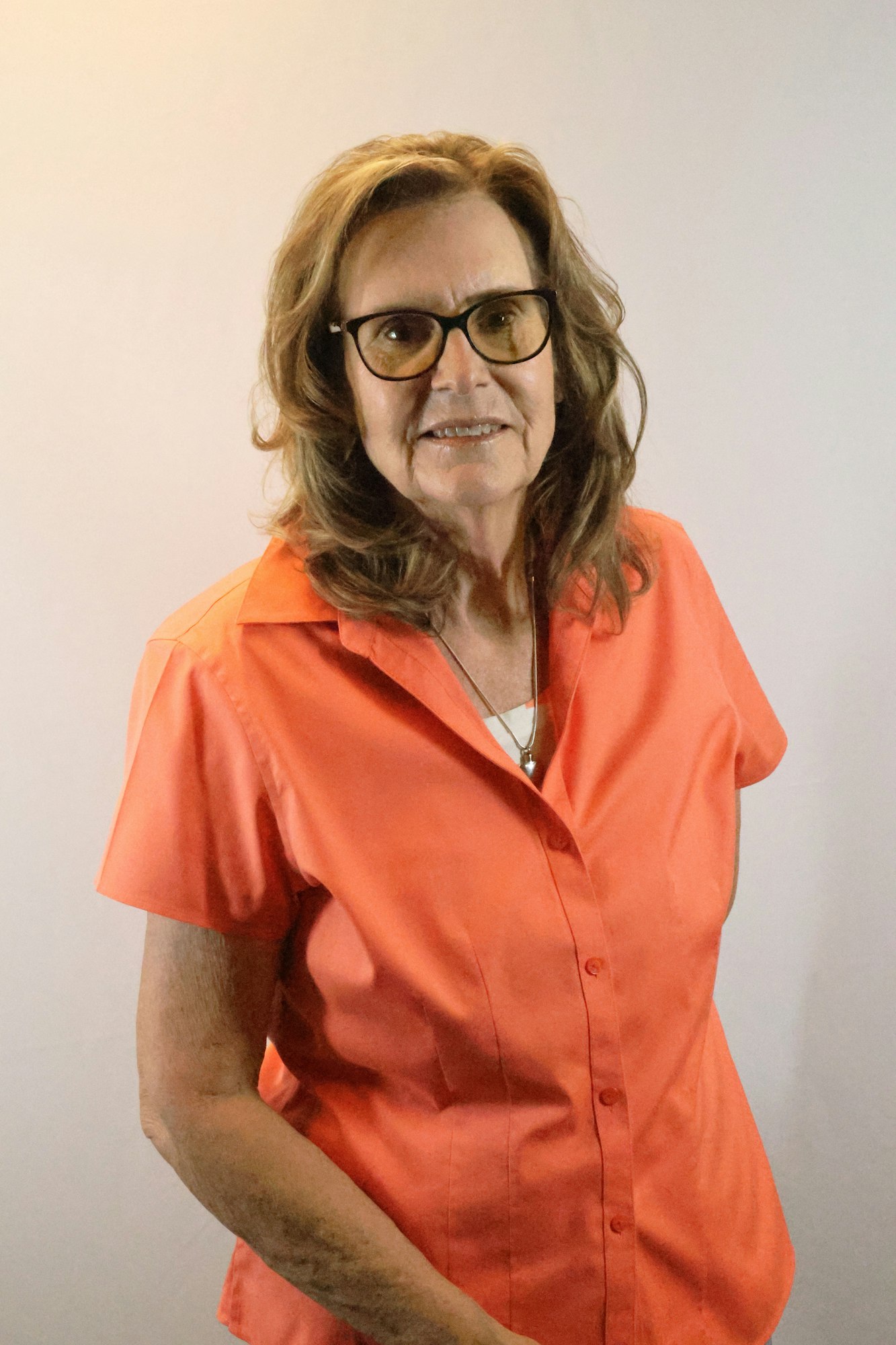 A person with glasses and long hair wearing an orange shirt, standing against a plain background.