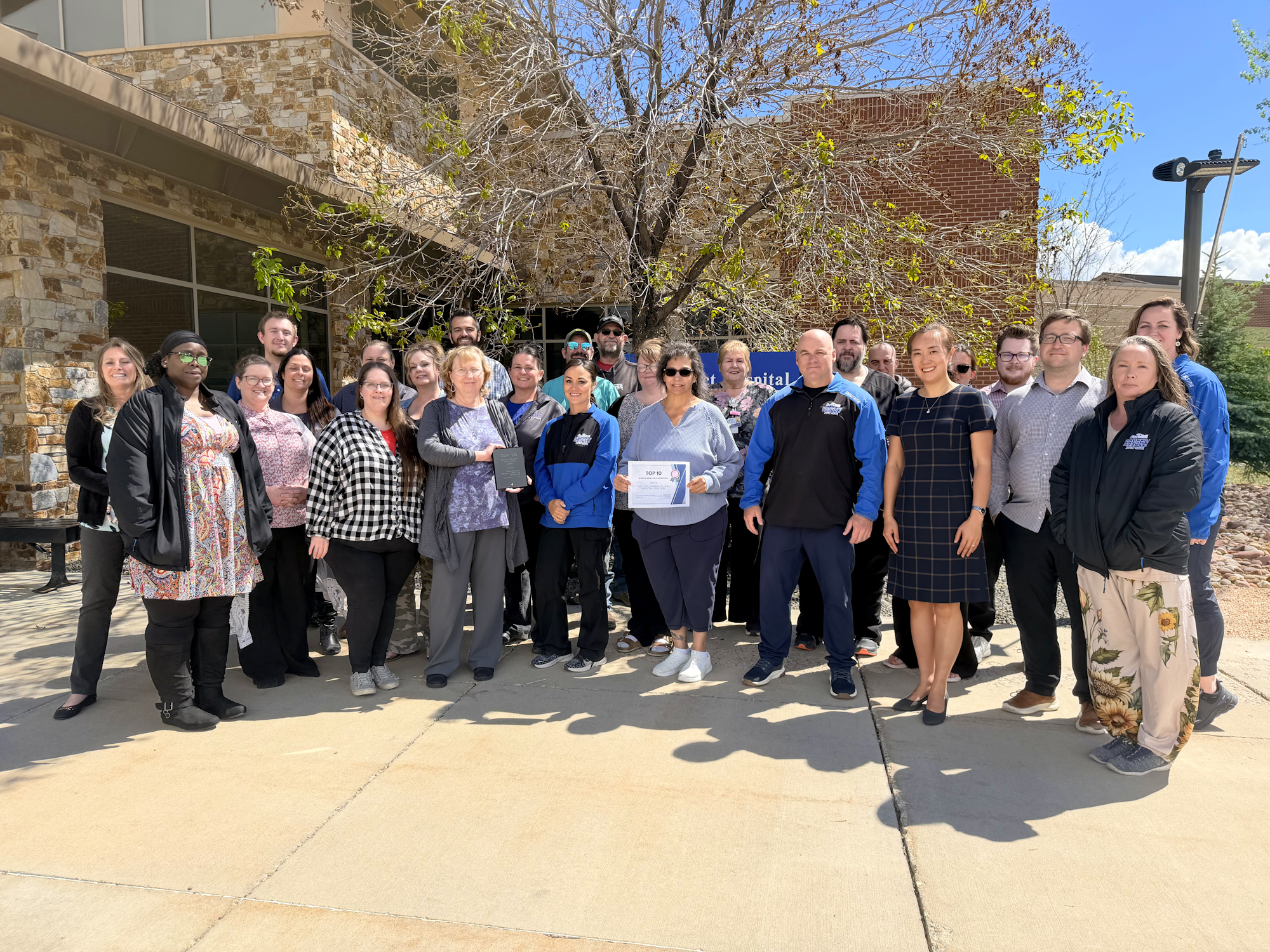 A group photo of a diverse team gathered outside a building, holding an award or certificate, smiling together.