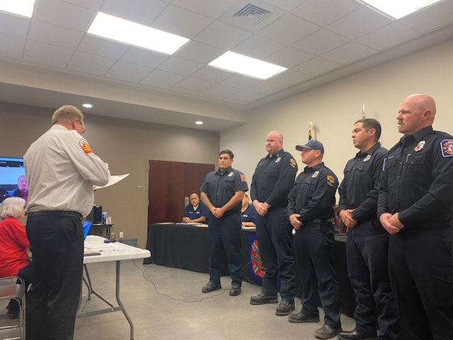 A group of firefighters standing, possibly during a ceremony or meeting, with an official speaking to them.