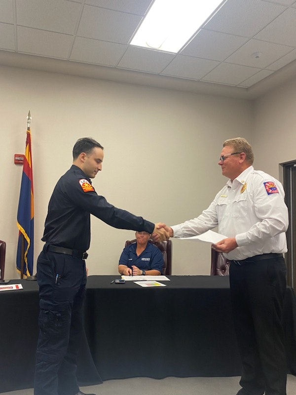Two individuals shaking hands, one in fire department uniform, other in formal attire, with a witness at a table, indoors.