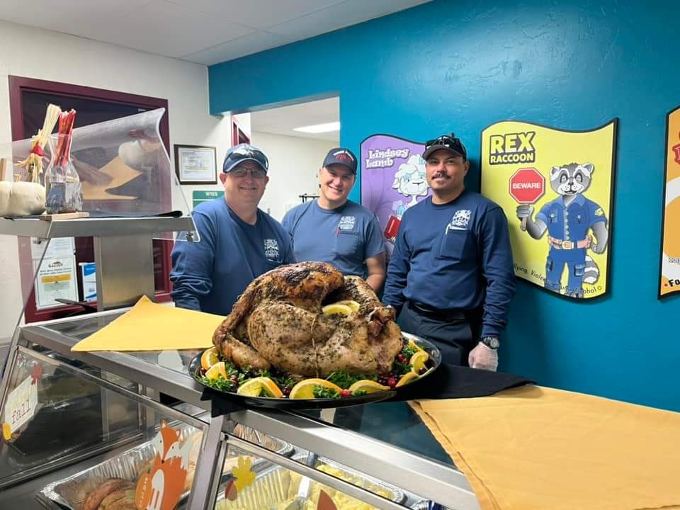 Three smiling individuals stand behind a beautifully presented roasted turkey, with colorful decorations in a lively setting.