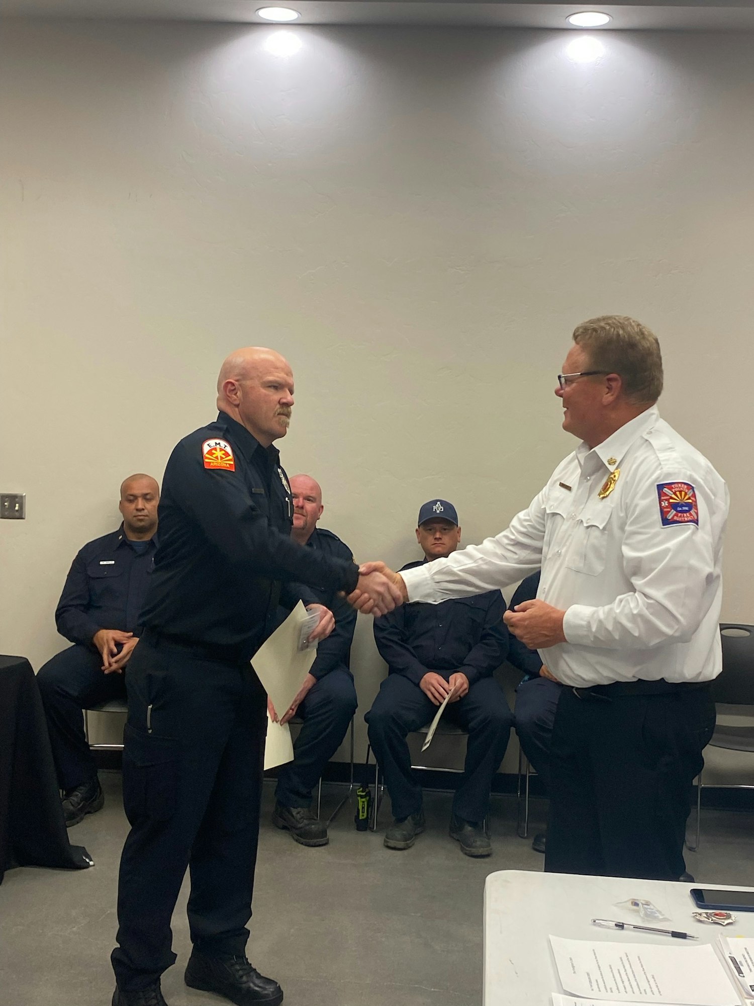 Two firefighters shaking hands, one receiving a document, with onlookers in a room.