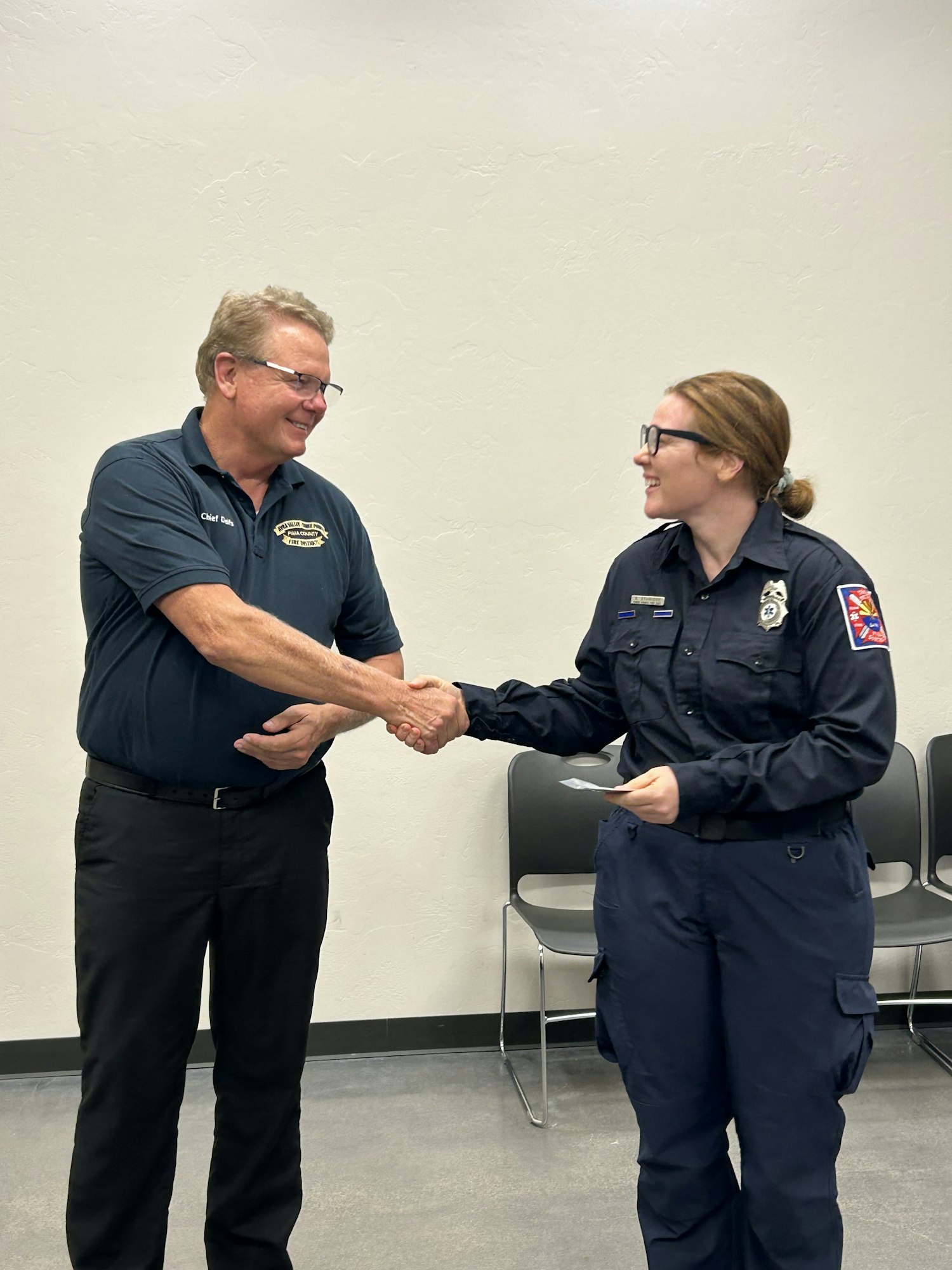 Two people shaking hands, one in a uniform, the other in a polo shirt, smiling in a room with chairs.