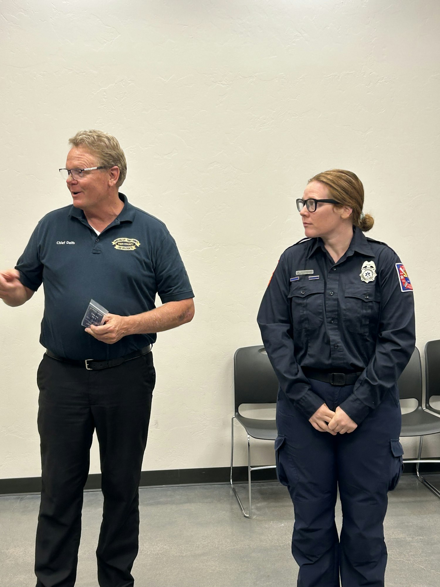 Two people standing; one in a uniform, the other in a polo shirt with a badge. There are chairs in the background.