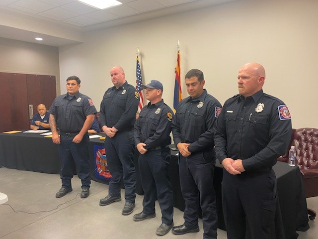 Five men in uniform, likely firefighters or EMS, standing in a room with an American flag and another person seated in the background.