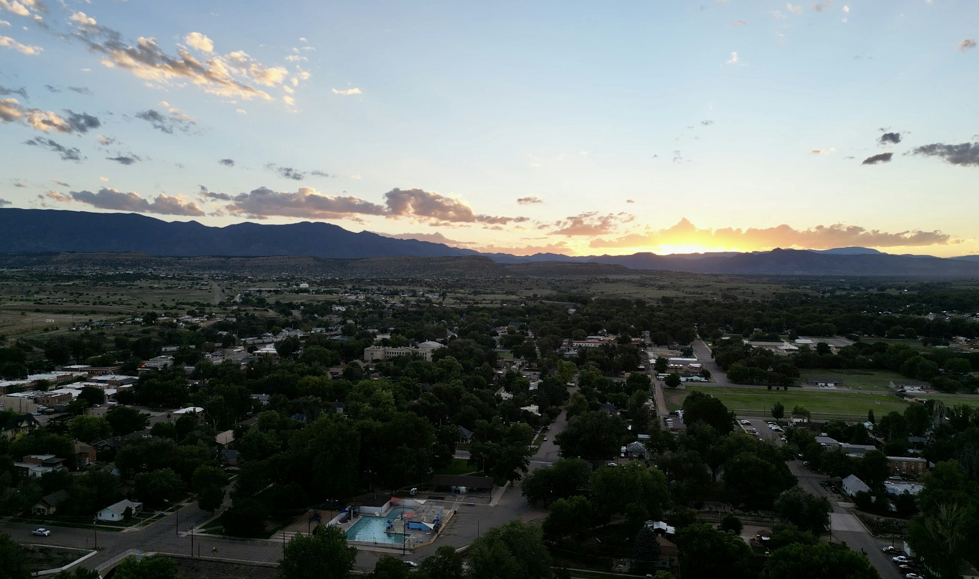 A sunset over a lush, residential area with mountains in the background and scattered clouds in the sky.