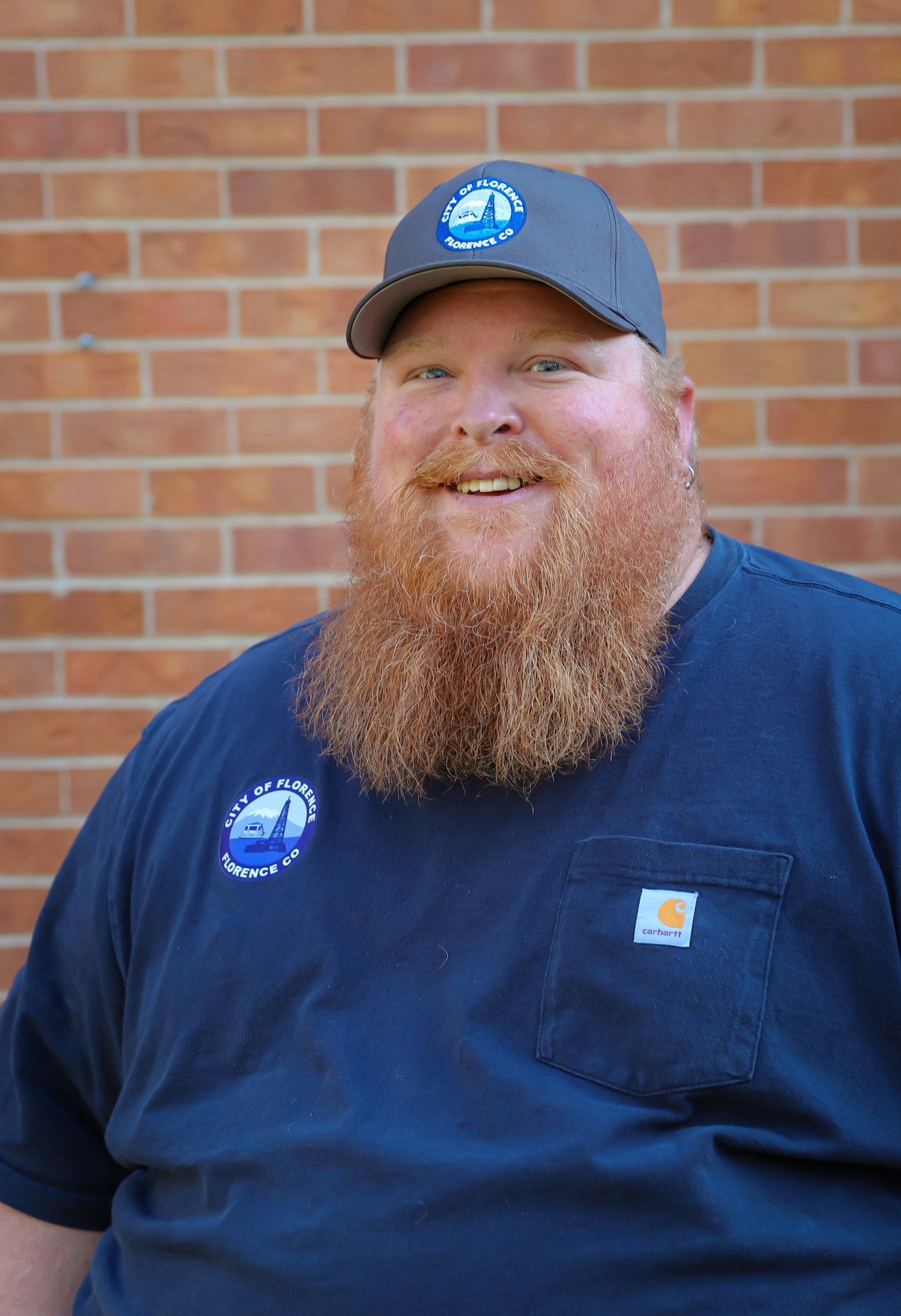 A smiling man with a thick beard, wearing a cap and blue shirt, stands in front of a brick wall.