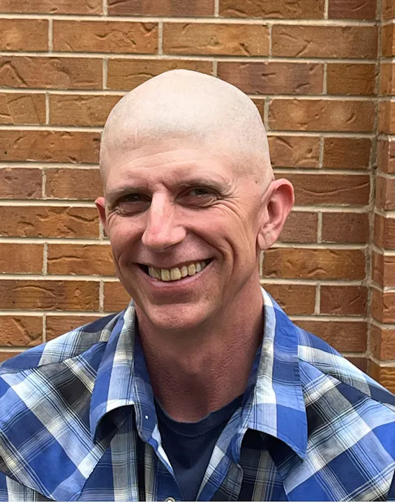 A smiling person with a shaved head, wearing a blue plaid shirt, poses in front of a brick wall.