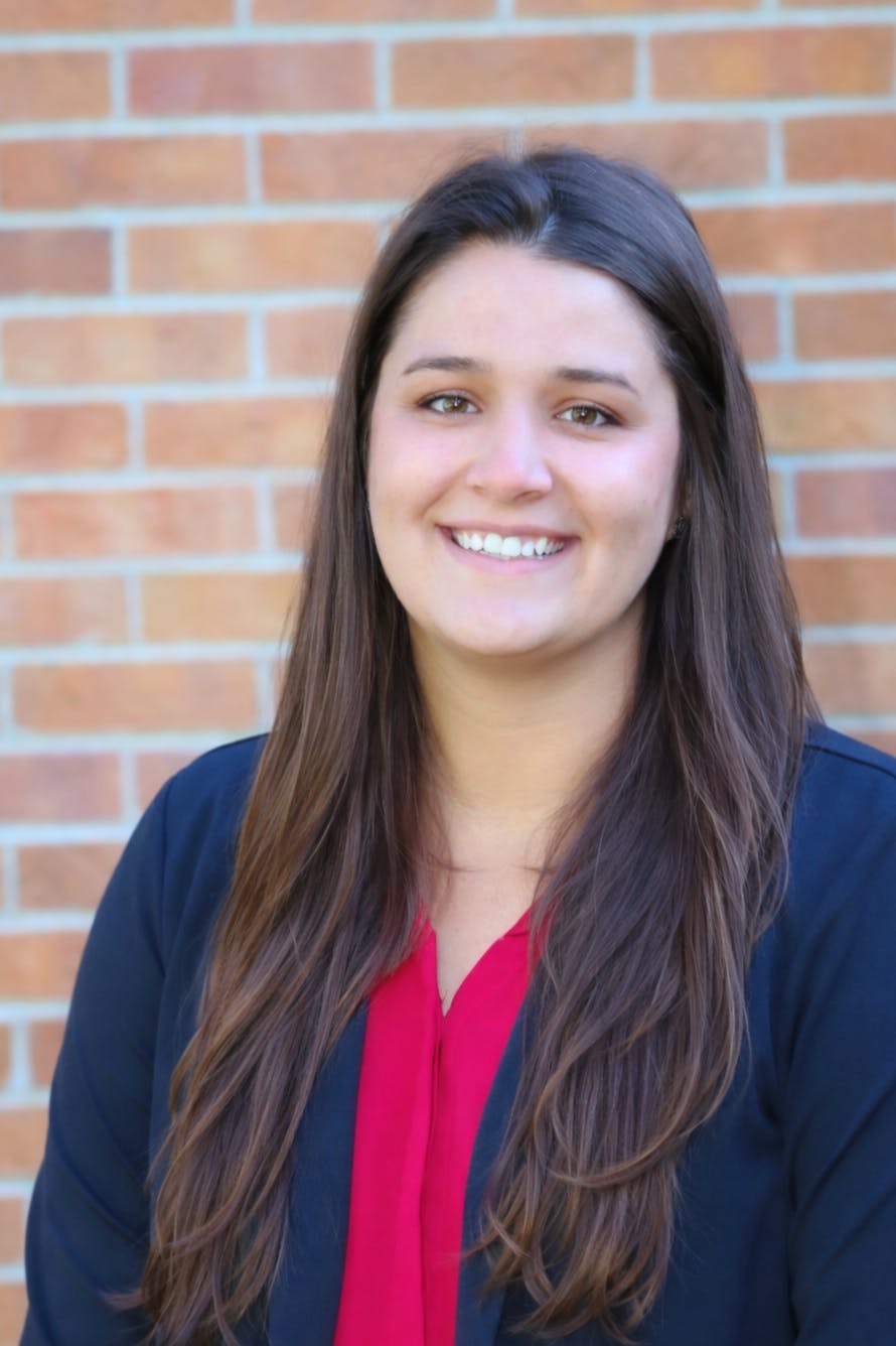 A woman with long hair smiles, wearing a red top and a navy jacket, against a brick background.
