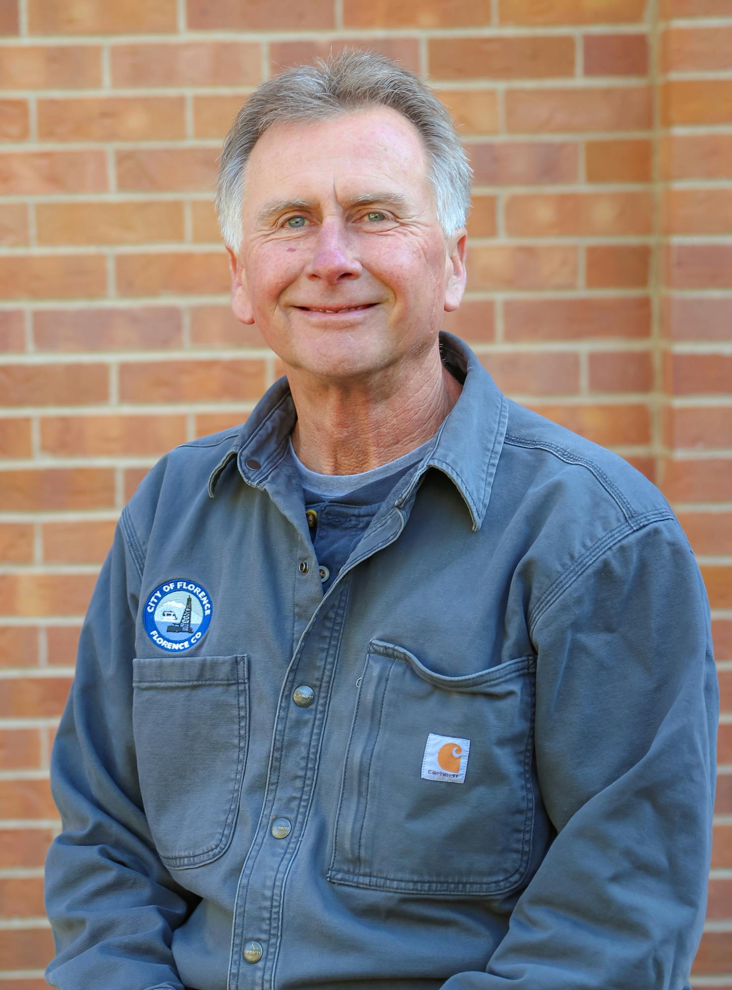 A smiling man in a gray work shirt sits on a rock against a brick wall, exuding a friendly demeanor.