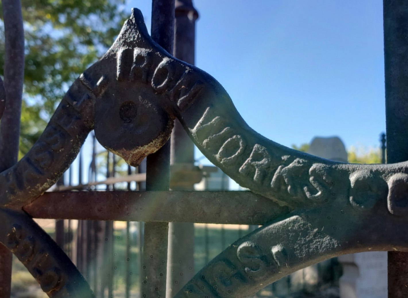 Close-up of a decorative iron fence with embossed text, blurred background, sunny day.