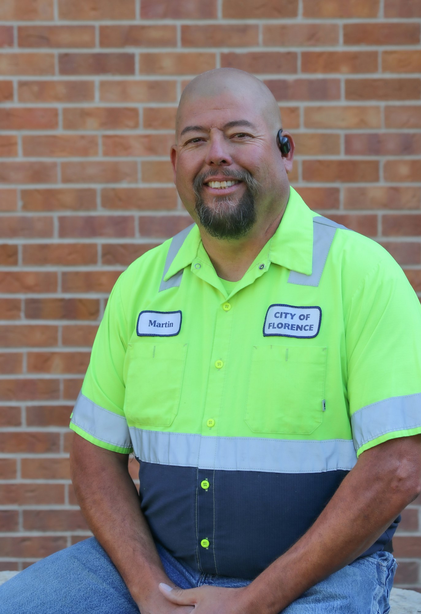 A smiling man in a bright yellow work shirt sits in front of a brick wall, displaying logos for the City of Florence.