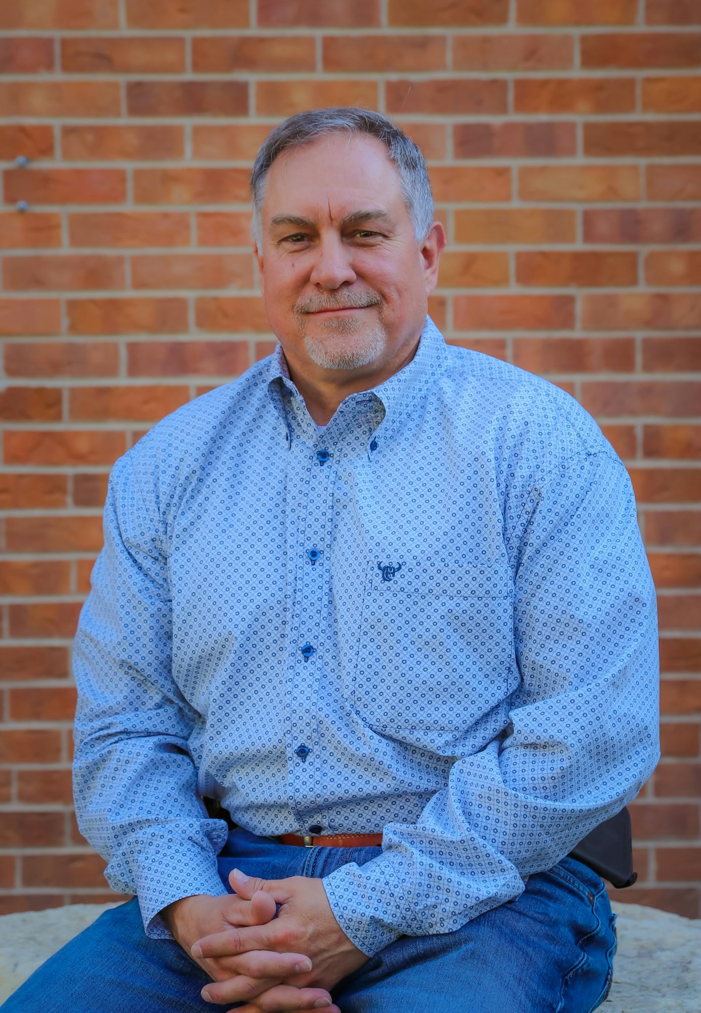 A man sits casually in front of a brick wall, wearing a patterned blue shirt and jeans, with a relaxed expression.
