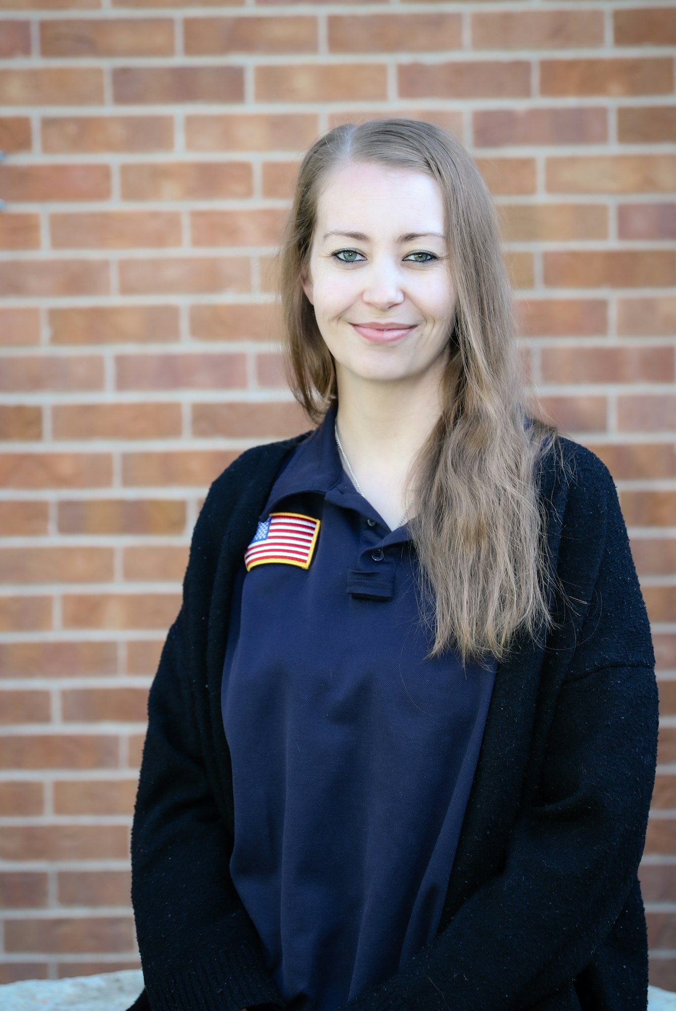 A woman with long hair is smiling, wearing a black cardigan over a navy shirt featuring an American flag, against a brick background.