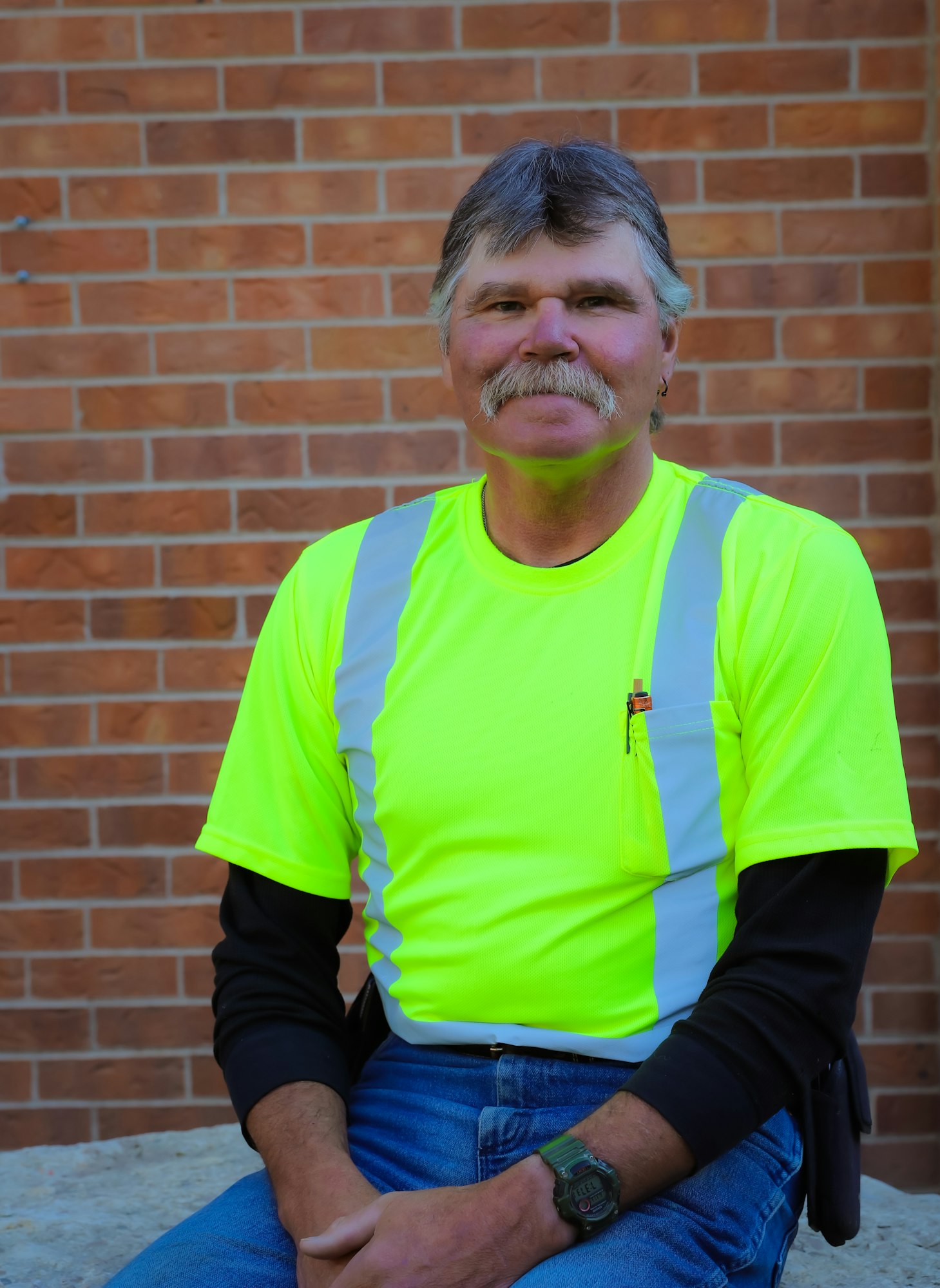 A man wearing a fluorescent yellow shirt and denim jeans sits against a brick wall, smiling confidently.