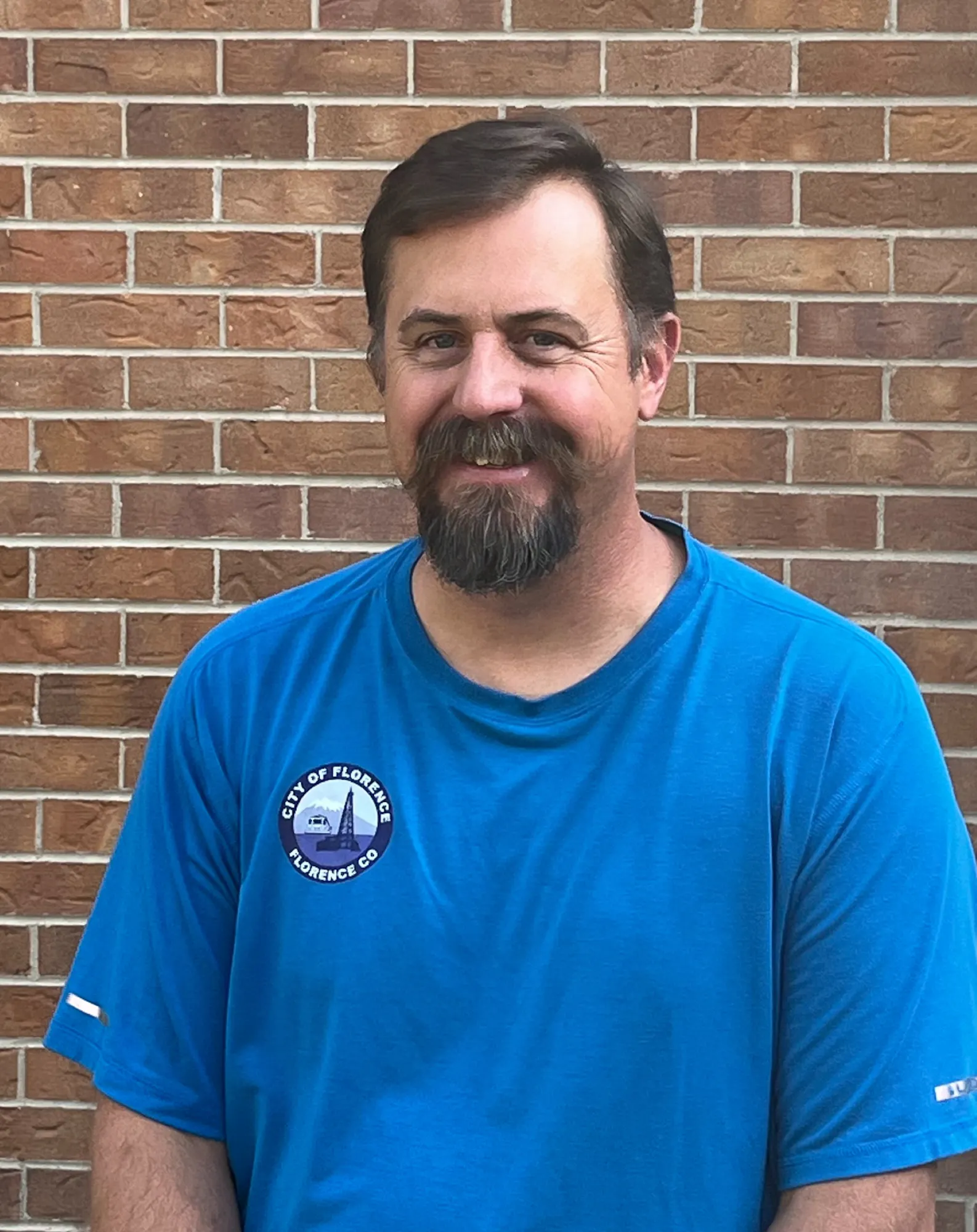 A man with a beard smiles, wearing a blue shirt with a city badge, standing in front of a brick wall.
