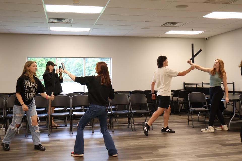 People in a room practicing with foam swords and taking photos, surrounded by chairs.