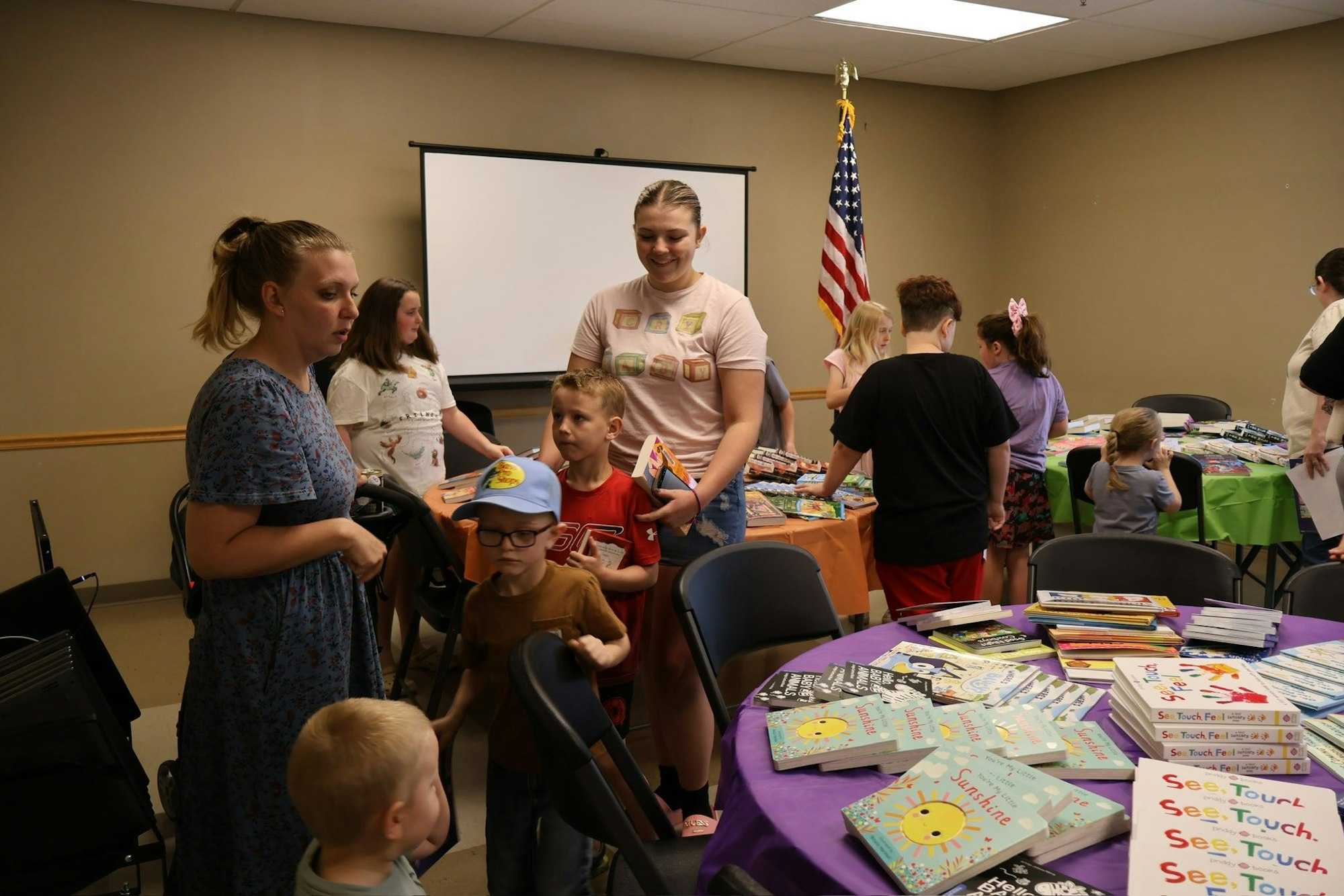 Kids and adults at a book fair or event, with tables of books and a U.S. flag in the background.