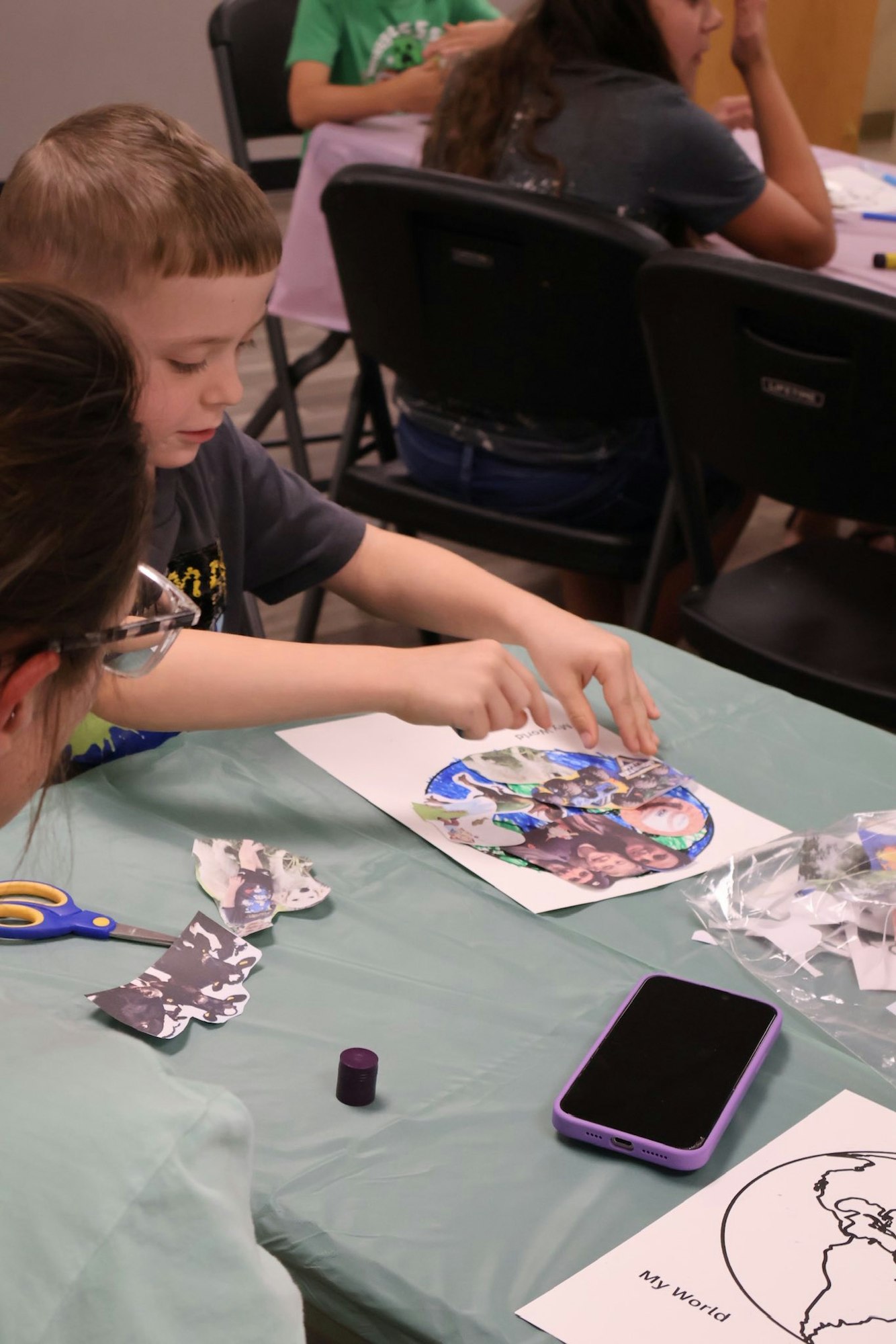 A child is engaging in arts and crafts, arranging a collage on paper. Nearby scissors, a phone, and artwork labeled "My World."