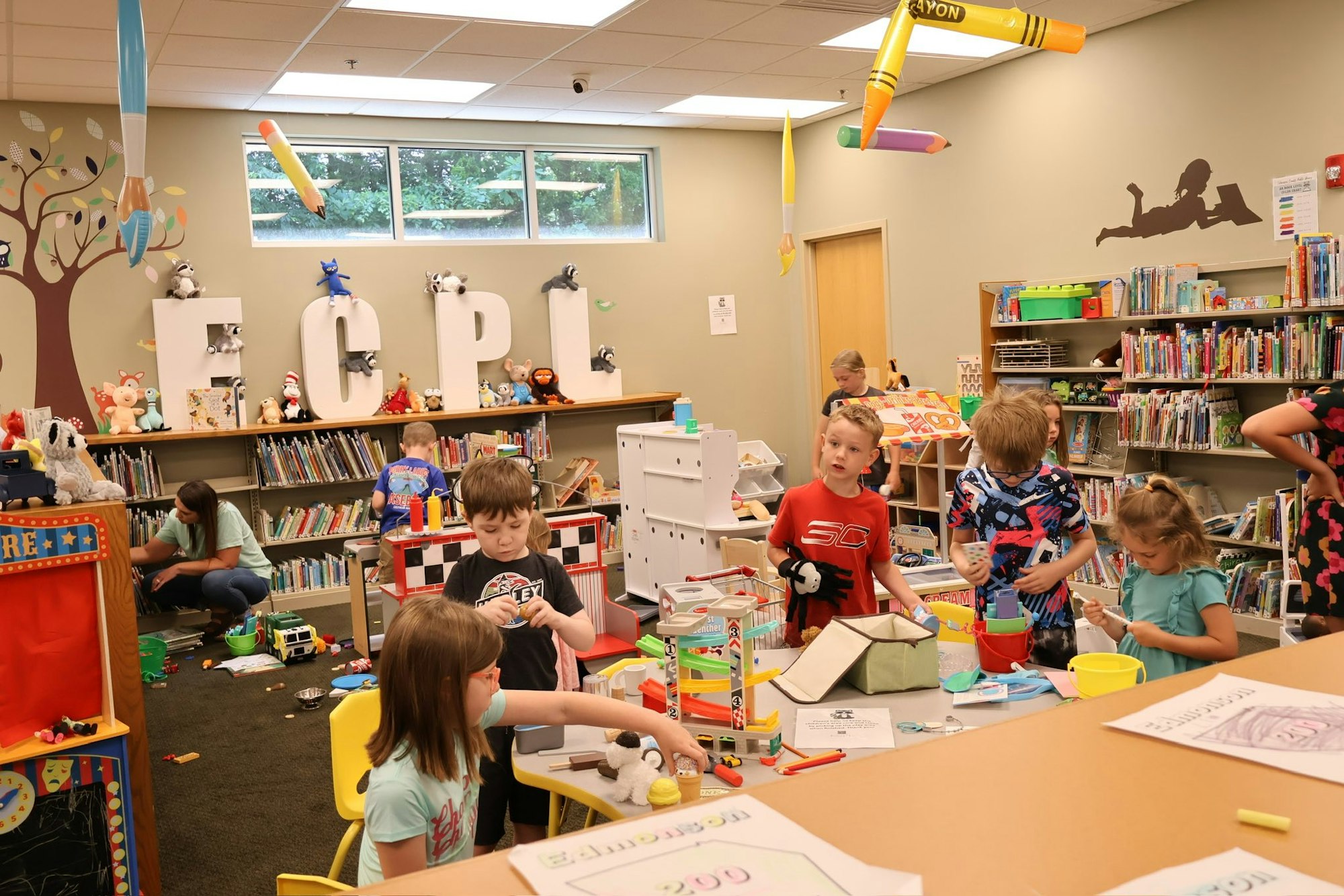 Children playing and reading in a colorful library room filled with books, toys, and large letter decorations.