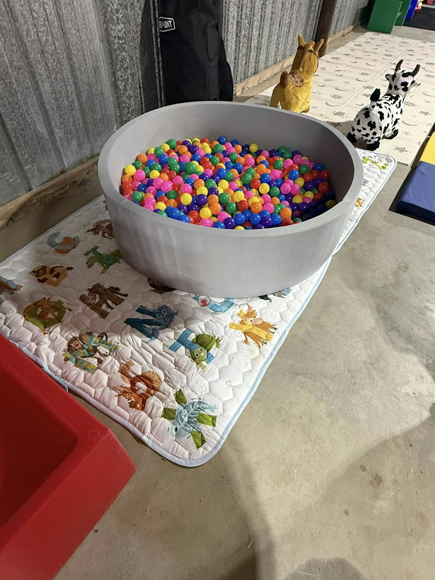 A gray ball pit with colorful balls sits on a quilted mat. Nearby are inflatable animal toys and a patterned floor mat.