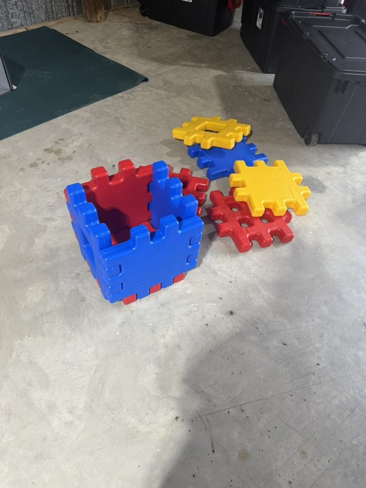 Large interlocking plastic blocks in red, blue, and yellow on a concrete floor, some assembled into a cube.