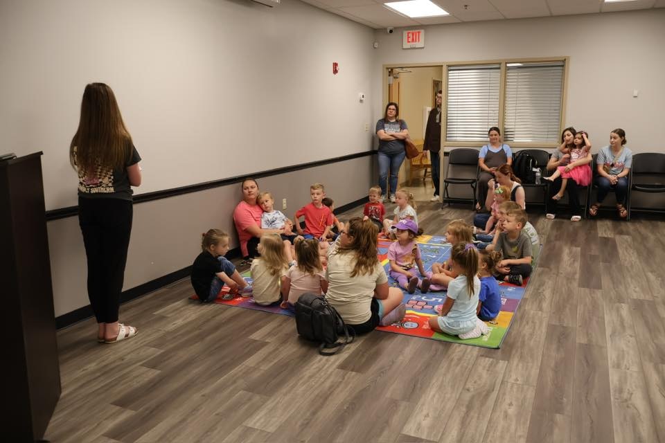 A group of children sits on a colorful mat in a room, while a woman stands at the front. Adults observe from chairs along the wall.