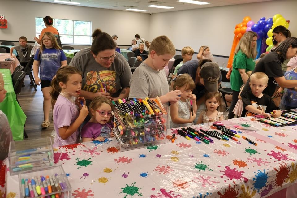 Children and adults at a colorful crafts table with markers and balloons in a room.