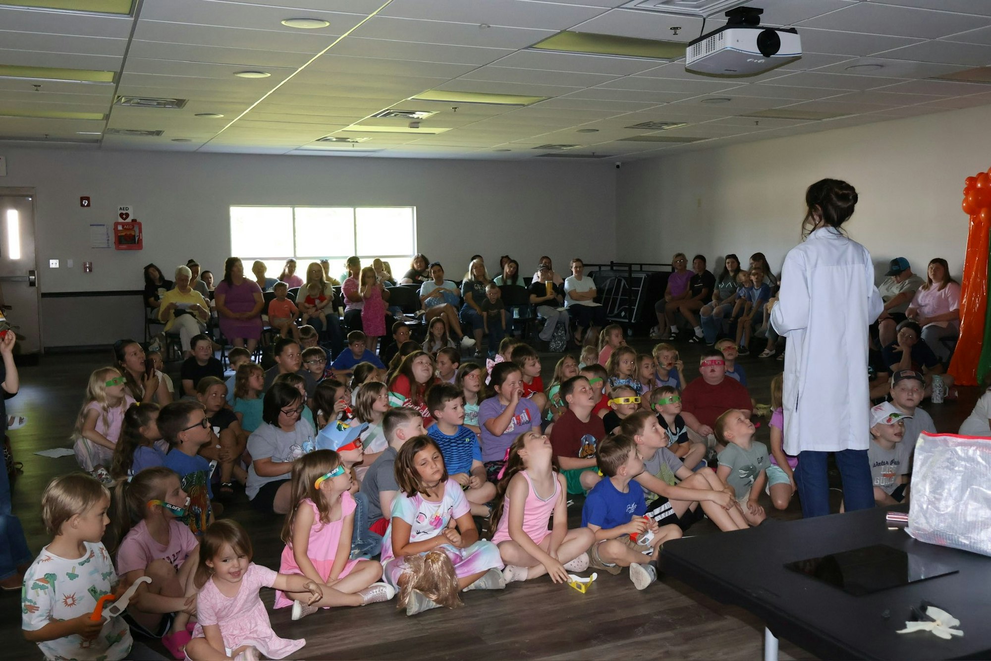 A person gives a presentation to a large group of children and adults in a classroom setting.