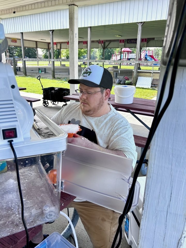 A person making shaved ice at an outdoor stand with tables, playground, and a machine visible.