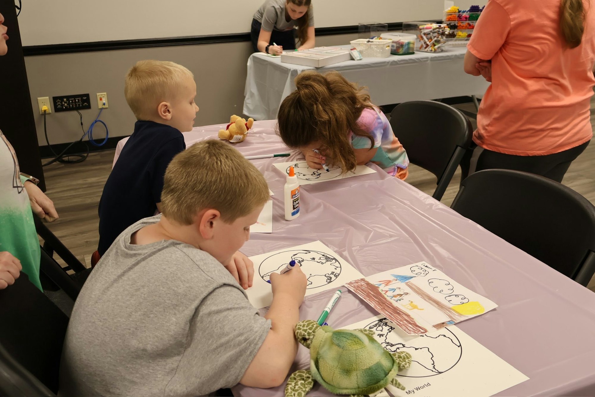 Children coloring world maps at a table with art supplies, including glue and markers.