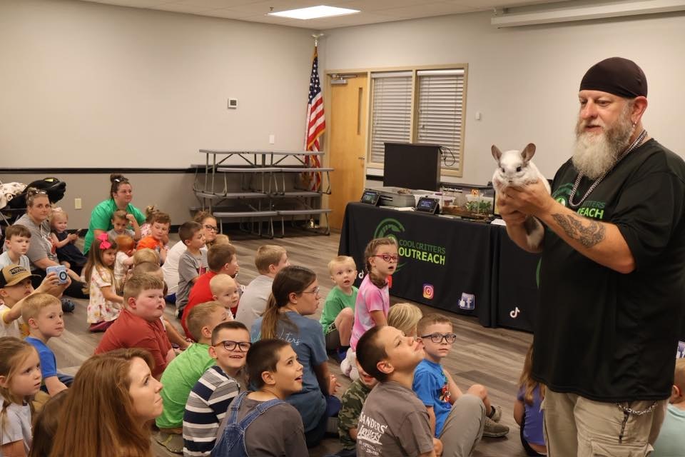 A man is holding a small animal in front of a group of attentive children seated on the floor.
